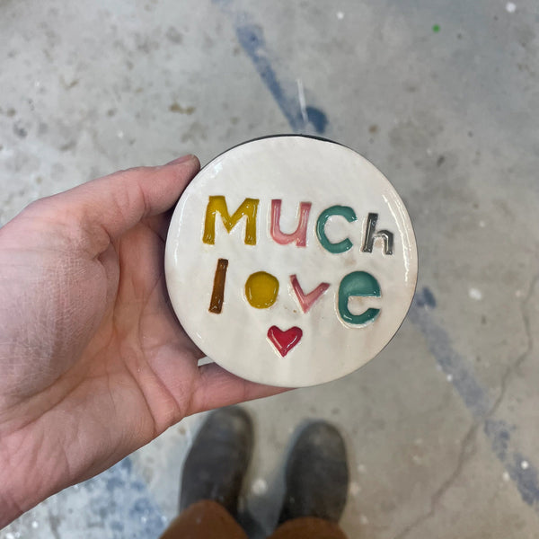 Round ceramic disc with 'Much Love' text held by a hand on a concrete floor.