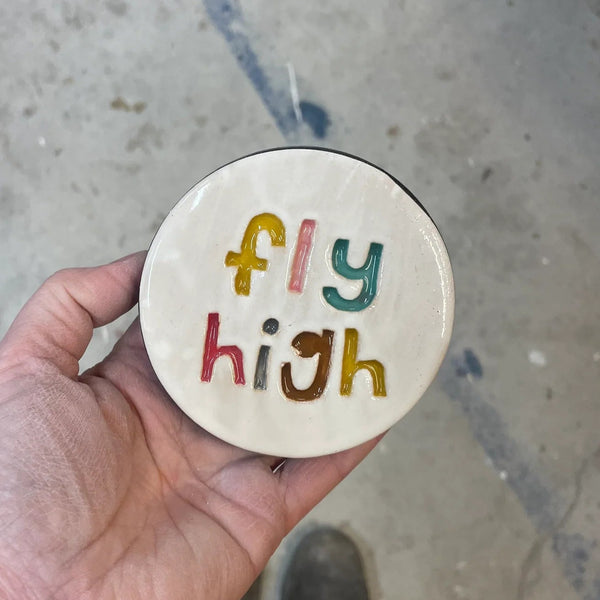 Round ceramic disk with 'fly high' text held by a hand on a concrete floor.