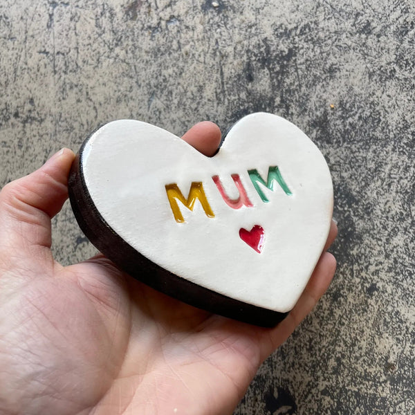 Heart-shaped ceramic with 'MUM' written on it held in a hand against a textured surface.