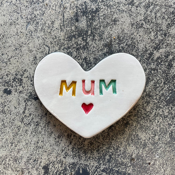 Heart-shaped ceramic with 'MUM' and a red heart on a gray surface