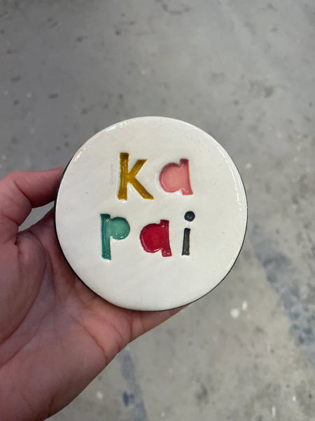Round ceramic item with colorful letters 'ka pai' held by a hand on a gray surface
