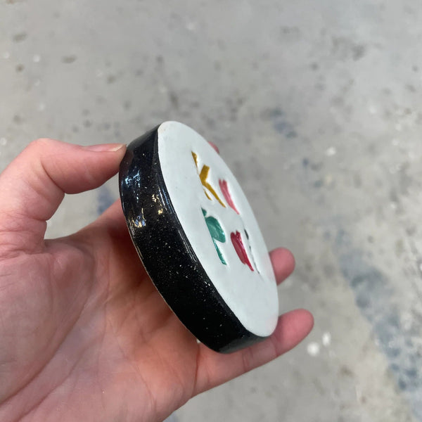 Hand holding a small round container with colorful designs on a gray pavement background